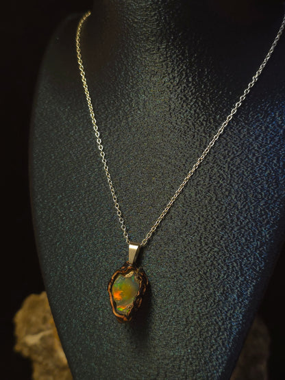 A silver chain necklace with a raw, irregular opal pendant displayed on a black textured mannequin bust against a dark background.