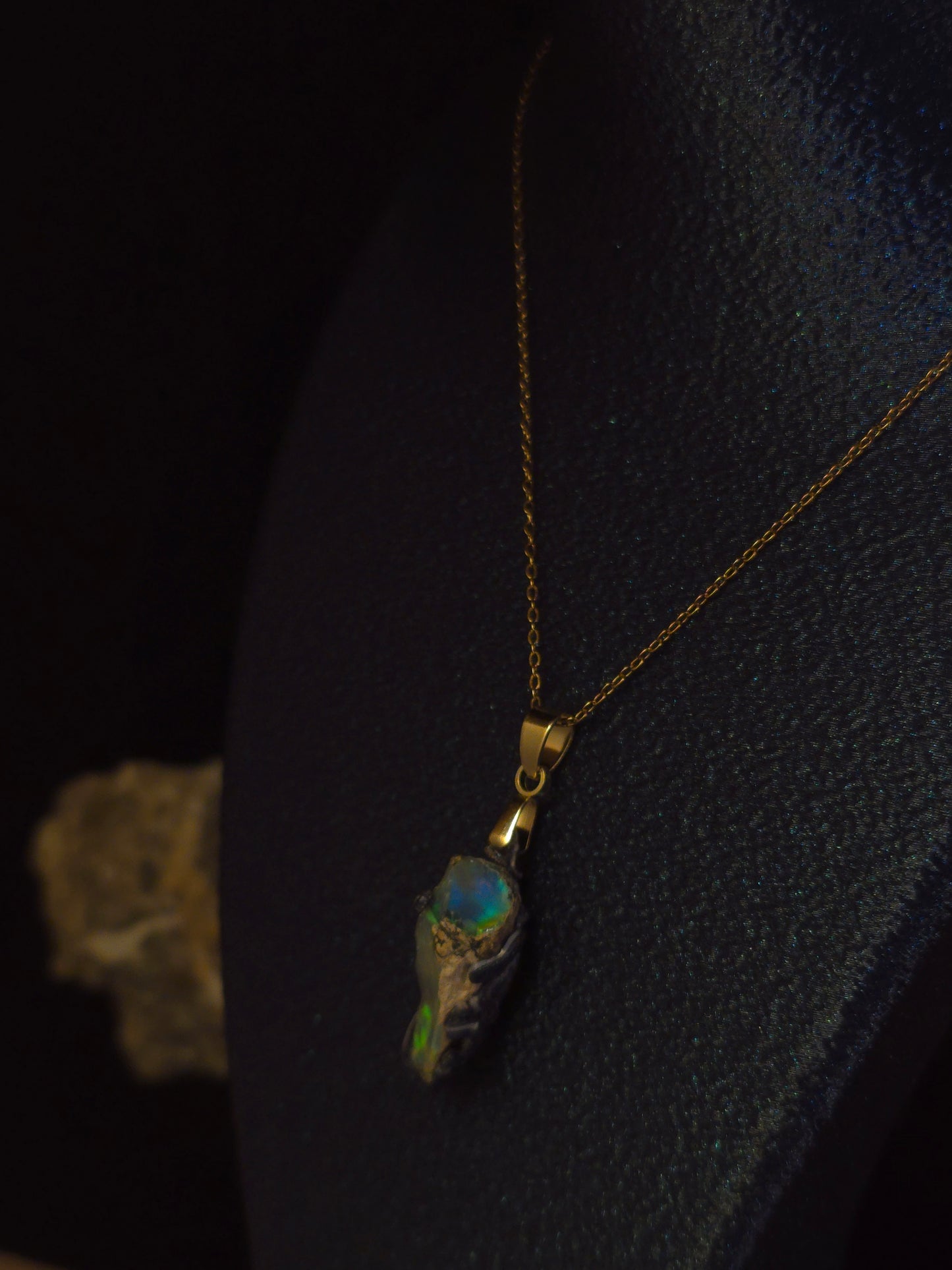 A gold chain necklace with a unique pendant featuring a textured gray and black base with vibrant iridescent green, blue, and orange opal inclusions, displayed on a dark textured bust.