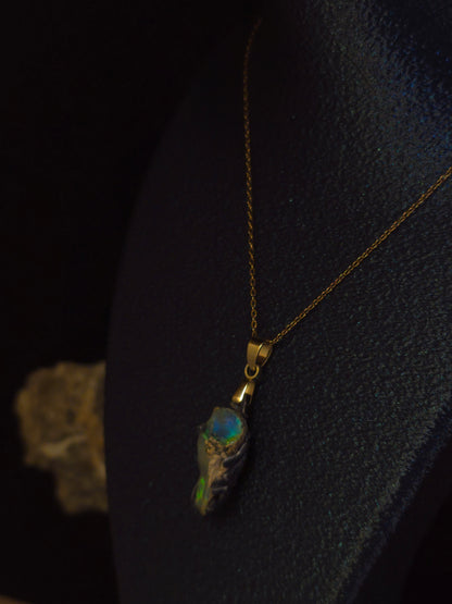 A gold chain necklace with a unique pendant featuring a textured gray and black base with vibrant iridescent green, blue, and orange opal inclusions, displayed on a dark textured bust.