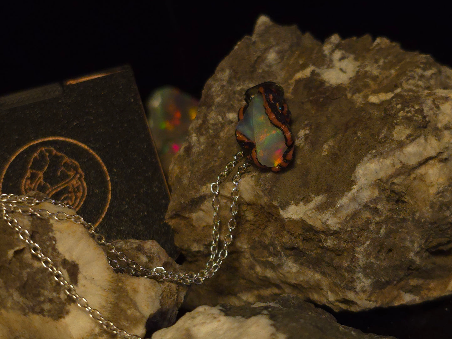 A silver chain necklace with a dark, irregularly shaped pendant featuring iridescent opal colors, displayed on textured rocks against a black background.