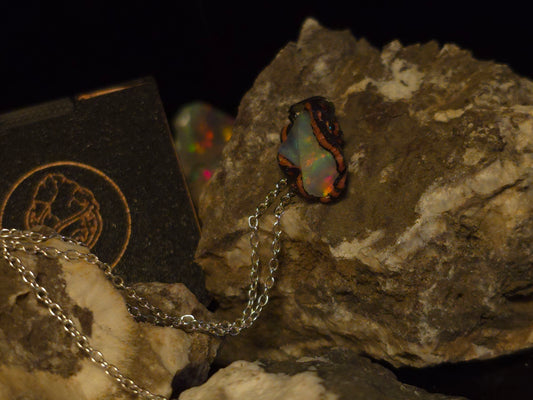 A silver chain necklace with a dark, irregularly shaped pendant featuring iridescent opal colors, displayed on textured rocks against a black background.
