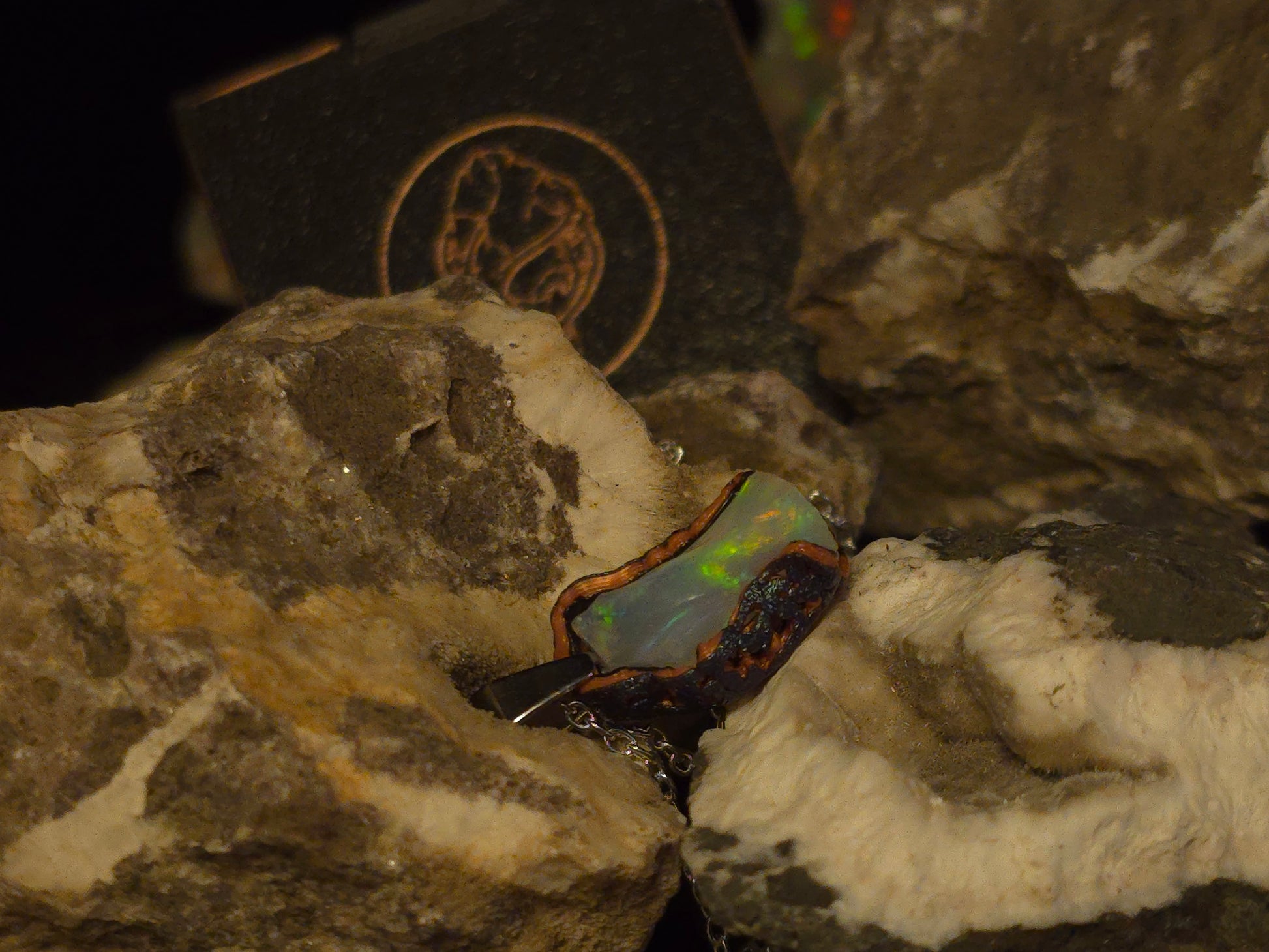A dark, irregularly shaped opal gemstone with iridescent green and blue flashes, set in a dark 3d printed bezel and displayed on textured white and brown rocks with a dark branded box in the background.