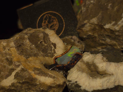 A dark, irregularly shaped opal gemstone with iridescent green and blue flashes, set in a dark 3d printed bezel and displayed on textured white and brown rocks with a dark branded box in the background.
