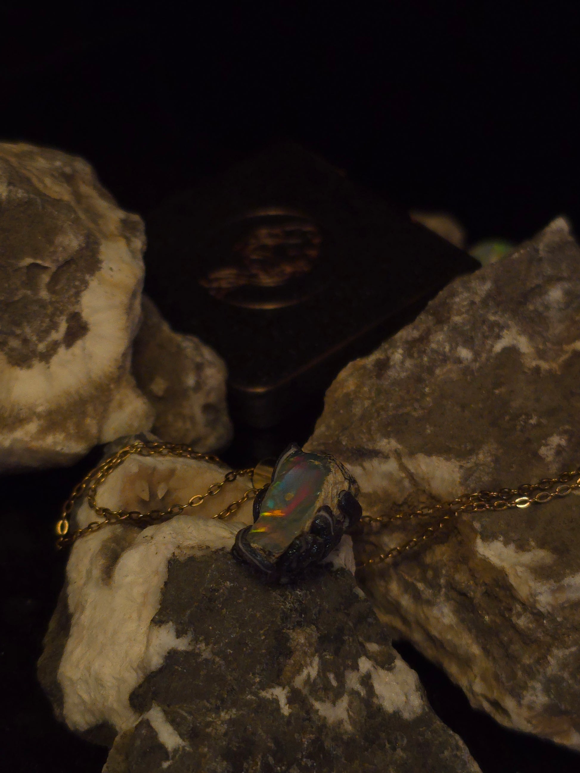 A gold chain necklace with a dark, iridescent opal-like pendant resting on rough natural rocks against a black background.