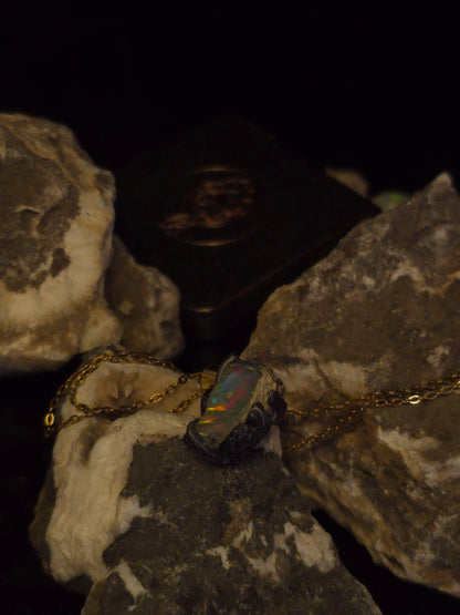A gold chain necklace with a dark, iridescent opal-like pendant resting on rough natural rocks against a black background.