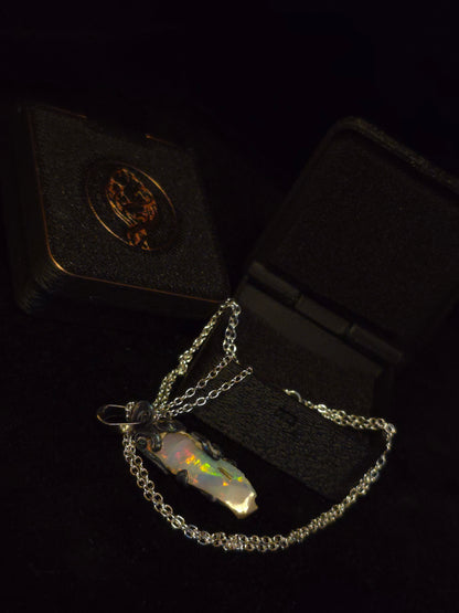 A silver chain necklace with a polished iridescent opal pendant, displayed on a black velvet jewelry box with a gold emblem.