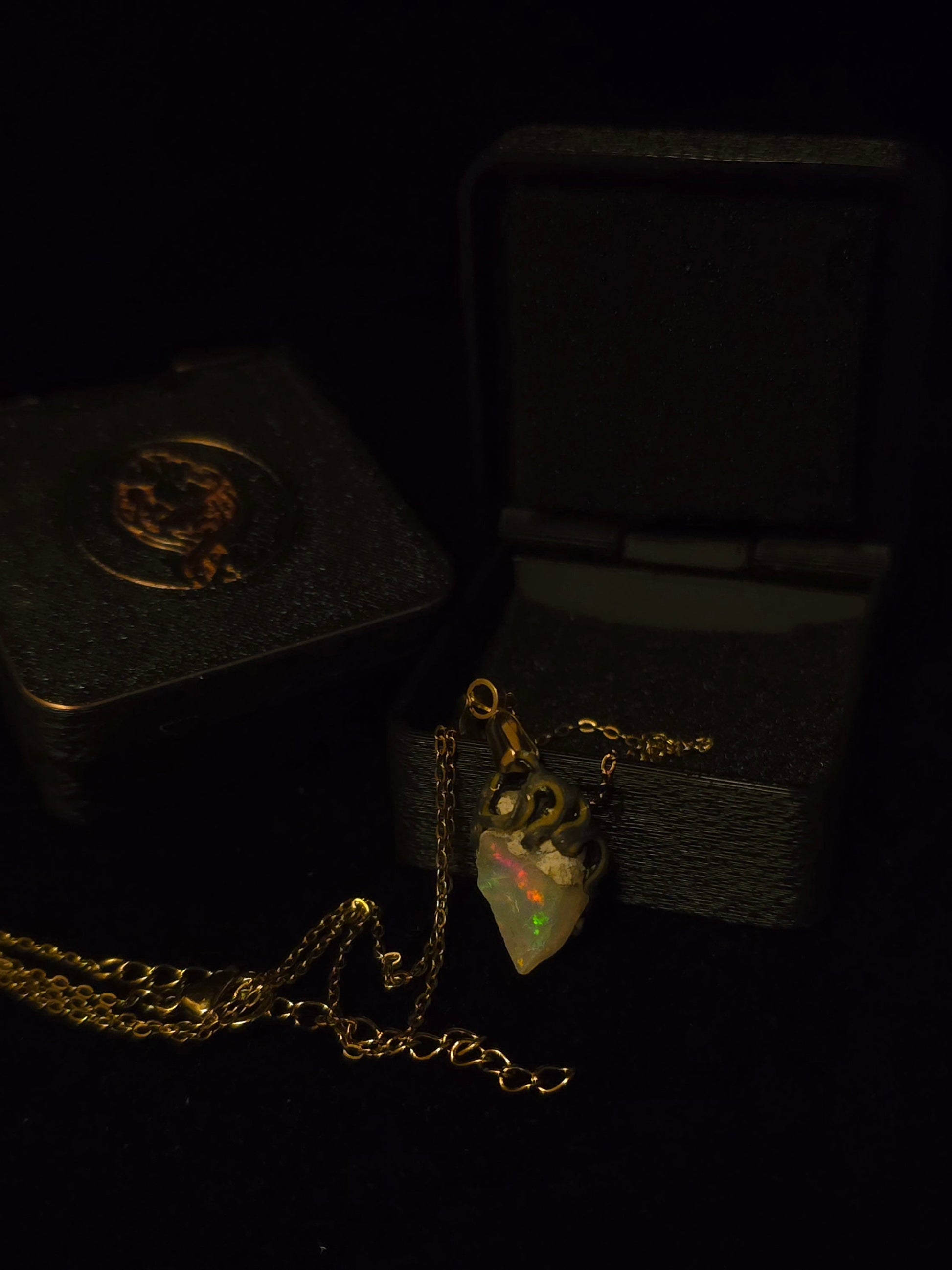 A gold pendant necklace with a teardrop-shaped opal gemstone, displayed next to its textured black presentation box on a dark background.