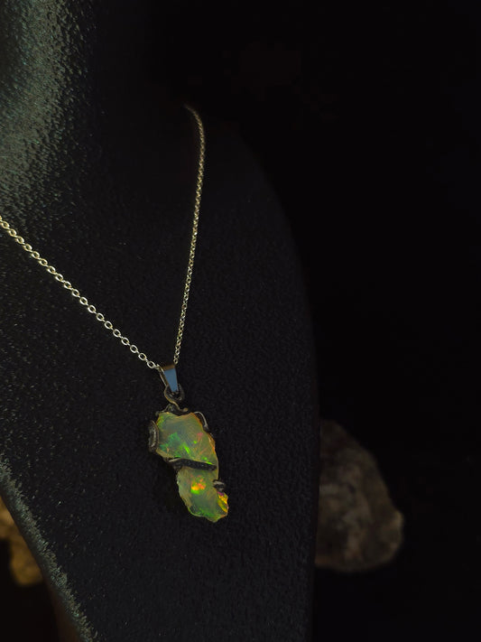 A delicate gold chain necklace featuring a raw, unpolished opal stone with vibrant green and yellow flashes, displayed on a black textured surface.