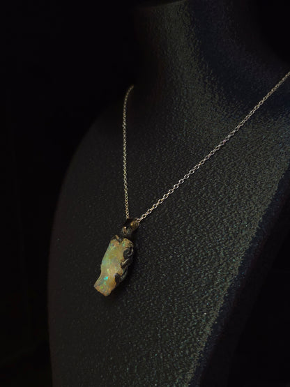 A necklace with a small irregular opal pendant on a fine silver chain, displayed on a black jewelry bust against a dark background.